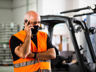 Serious adult male manager in uniform and protective mask standing with arms crossed near warehouse autoloader and having conversation on mobile phone - ADSF18796