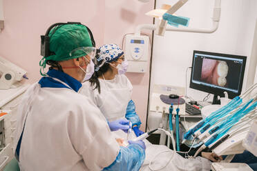 Rückenansicht eines nicht erkennbaren Arztes und einer Krankenschwester in Uniform in der Nähe eines Patienten und eines Desktop-Computers mit einem Zahnbild auf dem Bildschirm in einer Klinik - ADSF18829