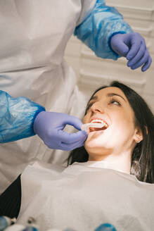 Von oben von einem nicht erkennbaren Kieferorthopäden in sterilen Handschuhen, der einer Frau in einer Klinik eine durchsichtige Zahnspange anlegt - ADSF18830