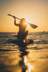 Unkenntlich Silhouette der Frau im Badeanzug sitzt auf SUP-Board in ruhigen Meer bei Sonnenuntergang im Sommer - ADSF18838