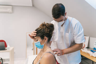 High angle of male physiotherapist massaging back of calm female patient in mask sitting on medical table in modern clinic - ADSF18839