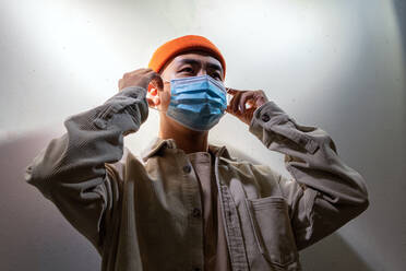 Concentrated Asian male in casual wear and protective face mask standing calmly in blurred gray room and looking away - ADSF18860