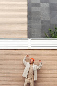 Überwältigter Mann in Freizeitkleidung, der schreit und vor Aufregung die Fäuste hebt, während er mit geschlossenen Augen vor einer gemauerten Wand steht - ADSF18867