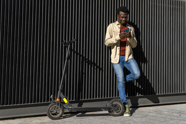 Glücklicher afroamerikanischer Mann, der eine SMS auf einem Mobiltelefon schreibt und auf einer Metallwand mit einem modernen Elektroroller entlang einer sonnigen Straße in der Stadt steht - ADSF18870