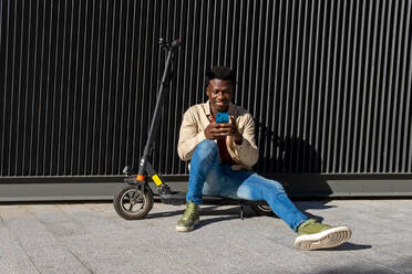 Happy African American male texting on mobile phone sitting on metal wall with modern electric scooter along sunny street in city - ADSF18871