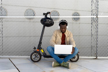 Black male sitting on parked electric scooter on street and messaging on social media via netbook while listening to music in headphones and enjoying weekend - ADSF18874