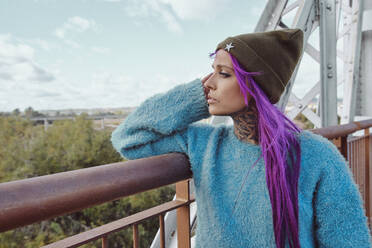 Trendy young female in pink hair leaning on metal fence and looking away - ADSF18900