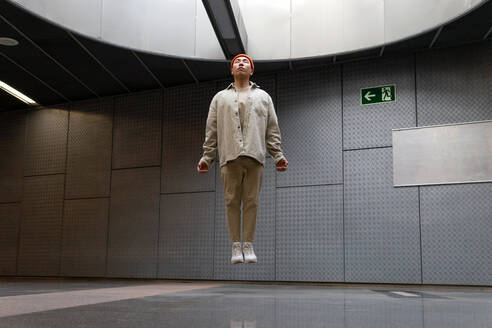 Ganzkörper fokussierter Stylist Asiatischer Mann in Freizeitkleidung schwebt mit geschlossenen Augen über dem Boden in einem Gebäudeflur - ADSF18923