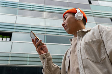 Von unten glücklicher asiatischer Mann in Freizeitkleidung und mit kabellosen Kopfhörern, der ein Videogespräch über sein Smartphone führt, während er vor einer modernen Eigentumswohnungsfassade steht - ADSF18927