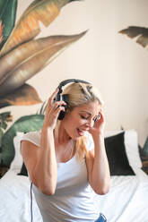 Dreamy female in headphones sitting on bed and enjoying songs with closed eyes and raised arms while entertaining at weekend - ADSF18954