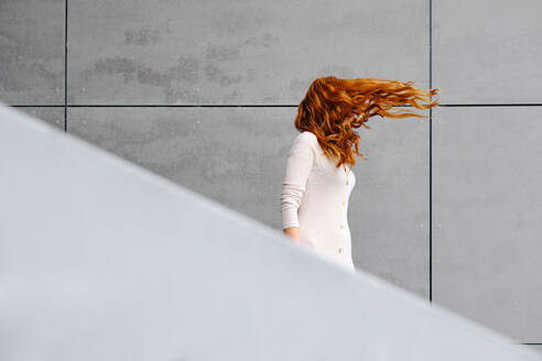 Seitenansicht einer nicht erkennbaren Frau mit langen roten Haaren, die im Wind flattern und an einer grauen Betonwand auf der Straße stehen - ADSF18958