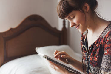 Lächelnde Frau, die im Schlafzimmer zu Hause Nachrichten auf ihrem digitalen Tablet sieht - MGRF00084