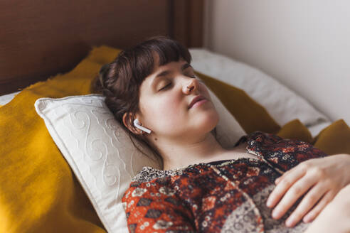 Junge Frau, die über drahtlose Kopfhörer eine geführte Meditation hört, während sie auf dem Bett liegt - MGRF00086