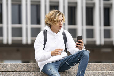 Junger Mann, der ein Mobiltelefon benutzt, während er auf einer Treppe sitzt - GGGF00450