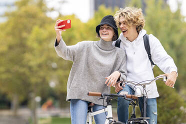 Happy woman taking selfie standing by male friend with bicycle in park - GGGF00452