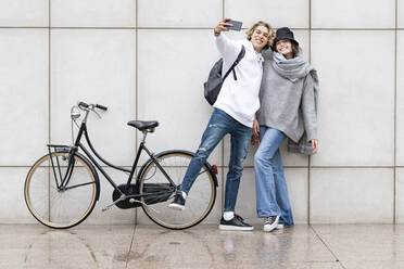 Mann, der ein Selfie macht, während seine Freundin mit dem Fahrrad auf dem Gehweg an der Wand steht - GGGF00454