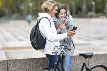 Junge Freunde mit Fahrrad lachen, während sie auf dem Gehweg ein Video über ihr Smartphone ansehen - GGGF00471