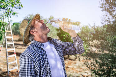 Seitenansicht eines durstigen Mannes mittleren Alters, der bei der Arbeit auf einer grünen Wiese frisches Wasser aus einer Plastikflasche trinkt - ADSF19050