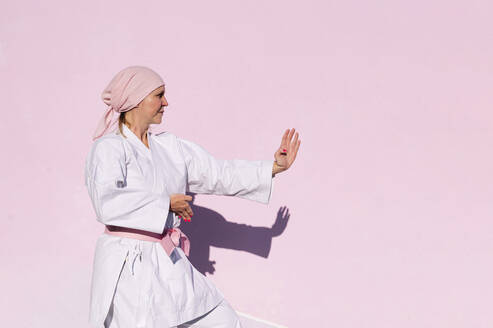 Seitenansicht der entschlossenen reifen Frau in rosa Kopfbedeckung und Gürtel kämpfen Karate in Krebs Kampf Konzept auf der Straße auf rosa Wand wegschauen - ADSF19055