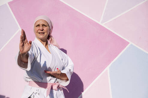 Entschlossen reife Frau in rosa Kopfbedeckung und Gürtel kämpfen Karate in Krebs Kampf Konzept auf der Straße auf rosa Wand wegschauen - ADSF19057