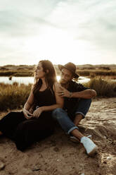 Young loving couple in casual clothing sitting together looking away on coast of reservoir surrounded by dry grass in meadow in sunlight - ADSF19061