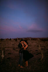 Full body side view of dreamy barefoot loving couple in casual clothing hugging with eyes closed while standing in meadow under vibrant blue pink sky at sunset - ADSF19065