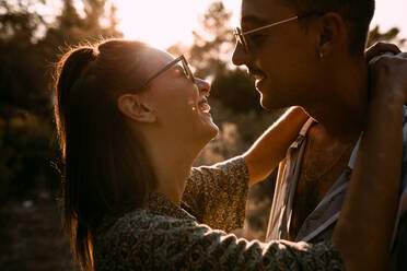 Side view of stylish couple in casual wear standing in nature and cuddling while looking at each other against sundown sky - ADSF19066