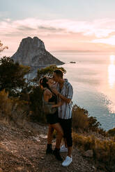 Seitenansicht eines leidenschaftlichen Paares, das sich umarmt und küsst, während es auf einem Hügel vor dem Hintergrund eines Sonnenuntergangs über dem Meer steht - ADSF19068