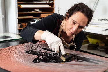 Reife Künstlerin in Schürze und Handschuhen, die eine gravierte Platte mit Tinte bedeckt, während sie ein Kunstwerk in einem professionellen Arbeitsraum erstellt - ADSF19093
