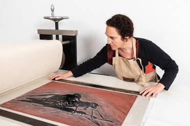 Fokussierter weiblicher Meister mittleren Alters, der ein Blatt Linoleum mit einem eingefärbten Ornament auf die Druckpresse legt, während er in der Linolschnitttechnik im Arbeitsatelier arbeitet - ADSF19096