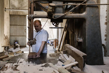 Seriöser männlicher Tischler, der eine Bandsäge benutzt und Holzbretter an einer Werkbank in einer chaotischen Werkstatt schneidet - ADSF19118