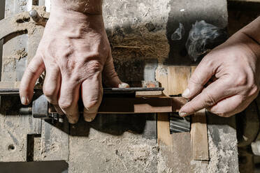 Männlicher Holzarbeiter steht an einer alten Werkbank und arbeitet mit Holzdetails in einer schmutzigen Werkstatt - ADSF19122