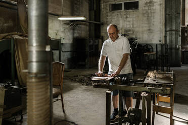 Entschlossener männlicher Holzarbeiter, der an einer alten Werkbank steht und mit Holzdetails in einer schmutzigen Werkstatt arbeitet - ADSF19125