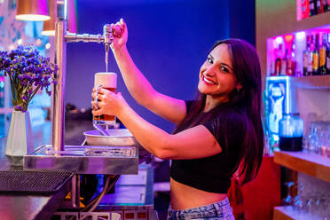 Seitenansicht einer Barkeeperin, die ein alkoholisches Getränk in ein Glas gießt, während sie in einer stilvollen Bar arbeitet - ADSF19141