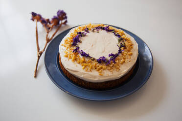 Appetitliche rustikale hausgemachte Torte mit weißer Sahne überzogen und mit Nüssen und essbaren Blumen dekoriert, serviert auf einem Teller auf einem weißen Tisch - ADSF19150