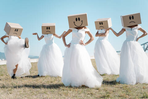 Full body of women wearing bridal dresses and boxes with happy and sad smileys on head standing in field stock photo