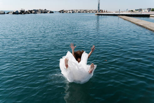 Unrecognizable young barefoot female in white wedding dress falling in rippling lake near pier looking at camera - ADSF19168