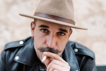 Cool handsome guy in with gorgeous mustache smoking cigarettes and looking at camera on blurred background - ADSF19174