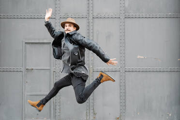 Full body of stylish thoughtful male with mustache in hat leaping and looking at camera near building with metal door and wall - ADSF19178