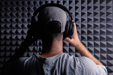 Back view of unrecognizable stylish male singer wearing headphones standing in recording studio on background of soundproof acoustic panel and looking at camera - ADSF19184