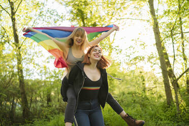 Glückliche Freundin, die eine Frau mit Regenbogenfahne huckepack nimmt, während sie im Wald steht - MTBF00789