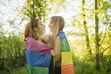 Happy lesbian couple standing wrapped in multi colored flag at forest - MTBF00792