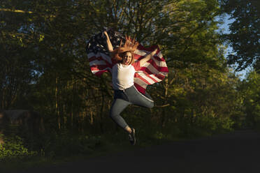 Side view of confident young ethnic lady in sports bra waving American flag  and looking at camera against modern glass skyscraper stock photo