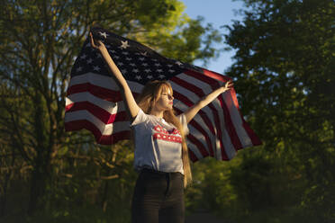 Blonde Frau hält amerikanische Flagge und steht mit geschlossenen Augen im Wald - MTBF00795