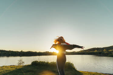 Carefree woman wearing jacket standing with arms outstretched against sky during sunset - MTBF00799