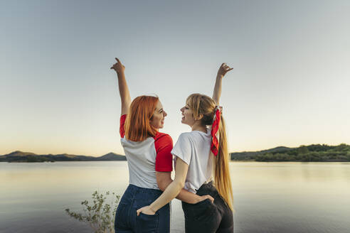 Smiling lesbian couple standing with hands in pockets against clear sky - MTBF00802