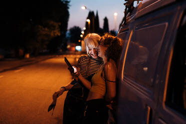 Female couple laughing with phone at night, leaning on van - CUF56614