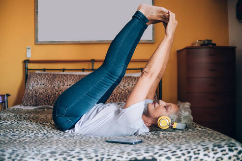 Woman stretching on bed, listening to headphones - CUF56618