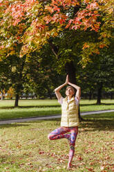 Ältere Frau, die auf einem Bein balanciert, übt Yoga im Herbstpark an einem sonnigen Tag - VYF00344