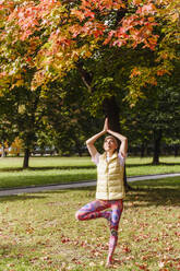Mature woman balancing on one leg, practicing yoga in autumn park on sunny day - VYF00344